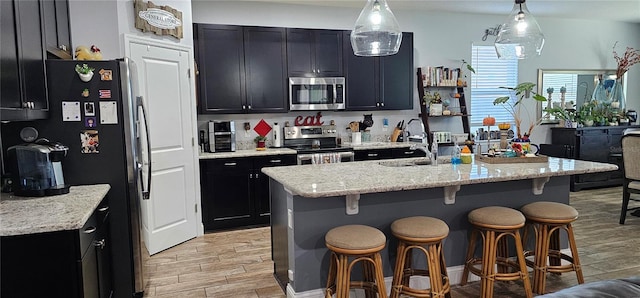 kitchen with light stone counters, light hardwood / wood-style flooring, pendant lighting, a center island with sink, and appliances with stainless steel finishes