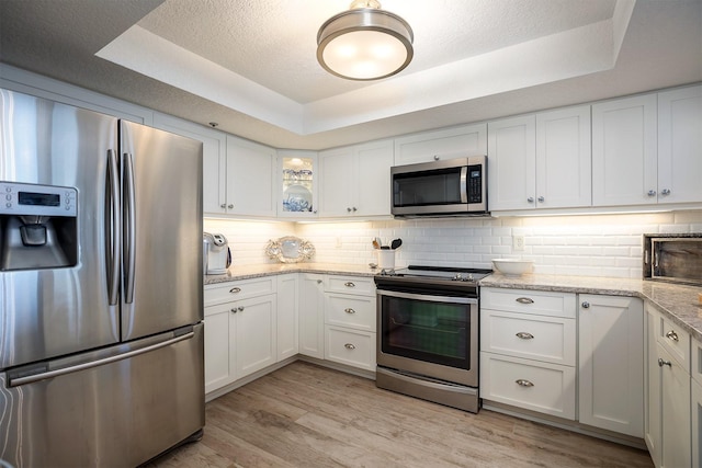 kitchen featuring appliances with stainless steel finishes, light stone counters, a tray ceiling, light hardwood / wood-style floors, and white cabinetry