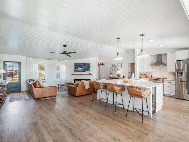 kitchen featuring a large island, wall chimney exhaust hood, stainless steel refrigerator with ice dispenser, decorative light fixtures, and white cabinets