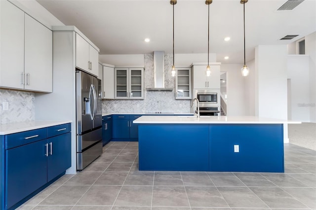 kitchen featuring wall chimney range hood, hanging light fixtures, blue cabinetry, an island with sink, and stainless steel appliances