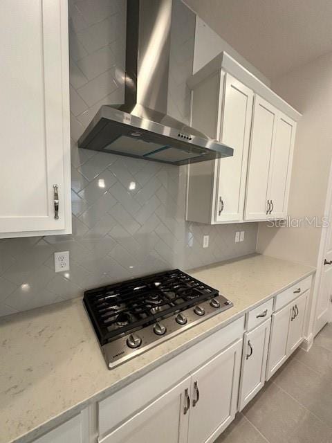 kitchen with light tile patterned flooring, stainless steel gas stovetop, white cabinetry, light stone counters, and wall chimney range hood