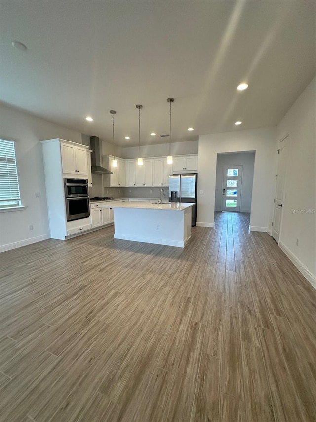 kitchen featuring decorative light fixtures, an island with sink, sink, white cabinets, and wall chimney range hood