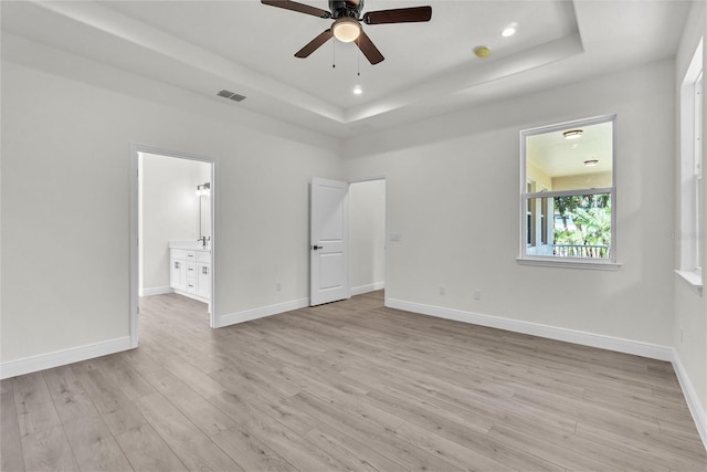 unfurnished bedroom featuring a raised ceiling, ensuite bath, ceiling fan, and light wood-type flooring