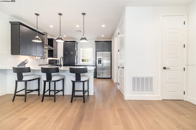 kitchen featuring wall chimney range hood, backsplash, hanging light fixtures, stainless steel fridge, and light hardwood / wood-style flooring