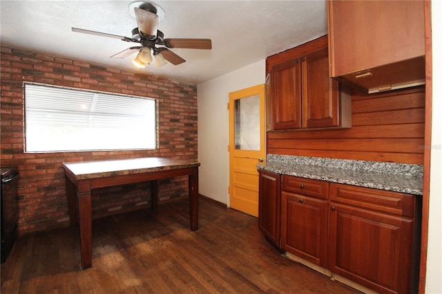 kitchen featuring dark wood-style floors, light stone counters, brown cabinets, ceiling fan, and brick wall