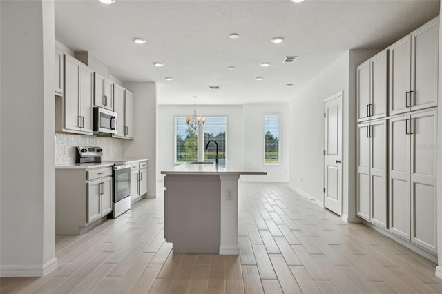 kitchen with sink, a kitchen island with sink, stainless steel appliances, tasteful backsplash, and decorative light fixtures