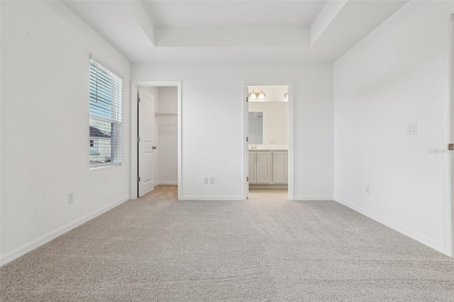 unfurnished bedroom featuring ensuite bath, a spacious closet, light carpet, a tray ceiling, and a closet