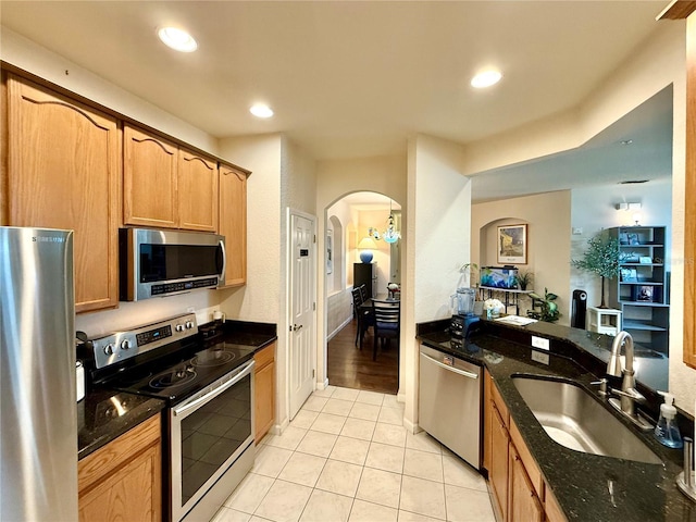 kitchen featuring arched walkways, dark stone countertops, stainless steel appliances, a sink, and light tile patterned flooring