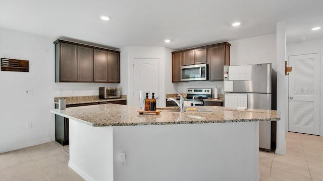 kitchen featuring a kitchen island with sink, dark brown cabinets, stainless steel appliances, and light stone counters