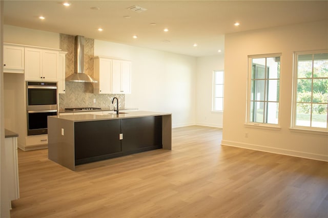 kitchen with wall chimney exhaust hood, a healthy amount of sunlight, an island with sink, and appliances with stainless steel finishes