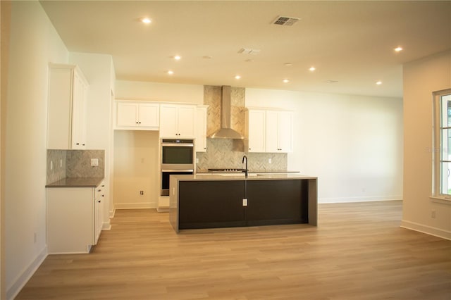 kitchen with double oven, wall chimney range hood, a center island with sink, white cabinets, and light hardwood / wood-style floors