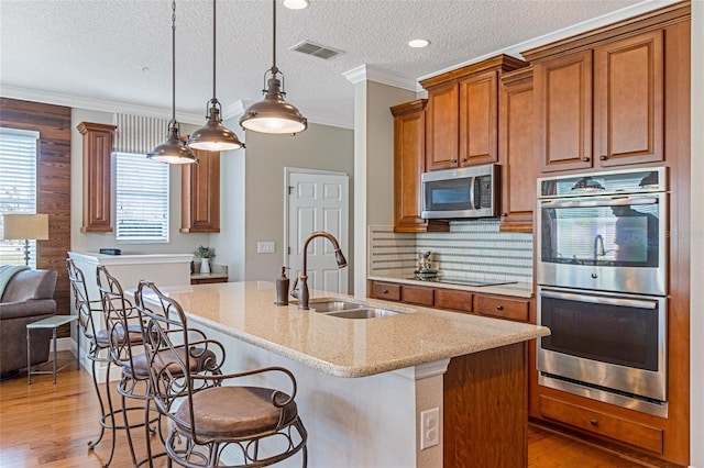 kitchen with light stone counters, stainless steel appliances, a kitchen island with sink, sink, and hanging light fixtures