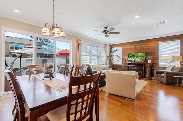 dining area with ornamental molding, ceiling fan with notable chandelier, a textured ceiling, wooden walls, and light hardwood / wood-style floors