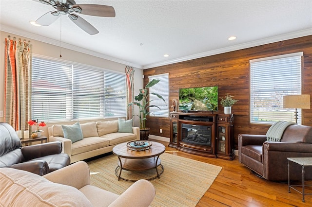 living room with ceiling fan, wood walls, light wood-type flooring, a textured ceiling, and ornamental molding