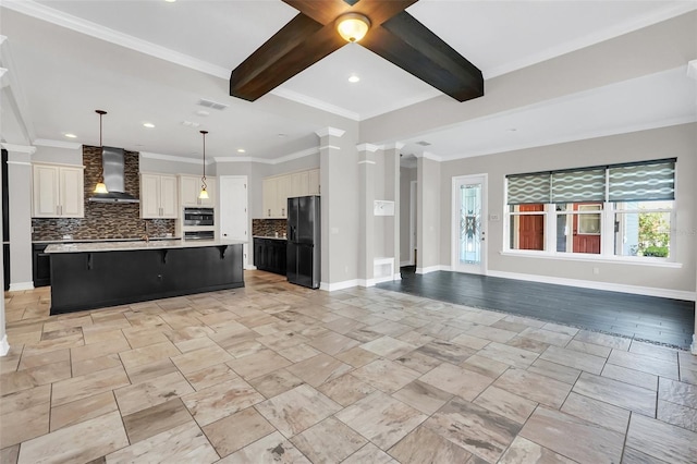 kitchen featuring a large island with sink, black fridge, wall chimney range hood, decorative light fixtures, and beam ceiling