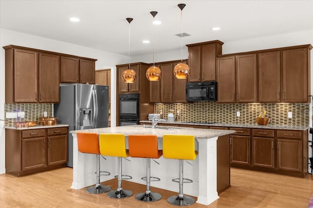 kitchen with light stone counters, black appliances, decorative light fixtures, a center island with sink, and a breakfast bar area