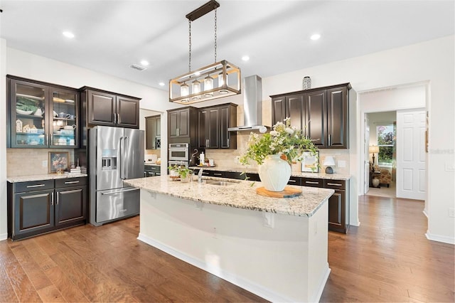 kitchen with wall chimney exhaust hood, decorative backsplash, an island with sink, dark brown cabinetry, and stainless steel appliances