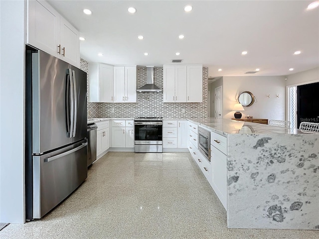 kitchen featuring kitchen peninsula, appliances with stainless steel finishes, light stone countertops, wall chimney range hood, and white cabinets