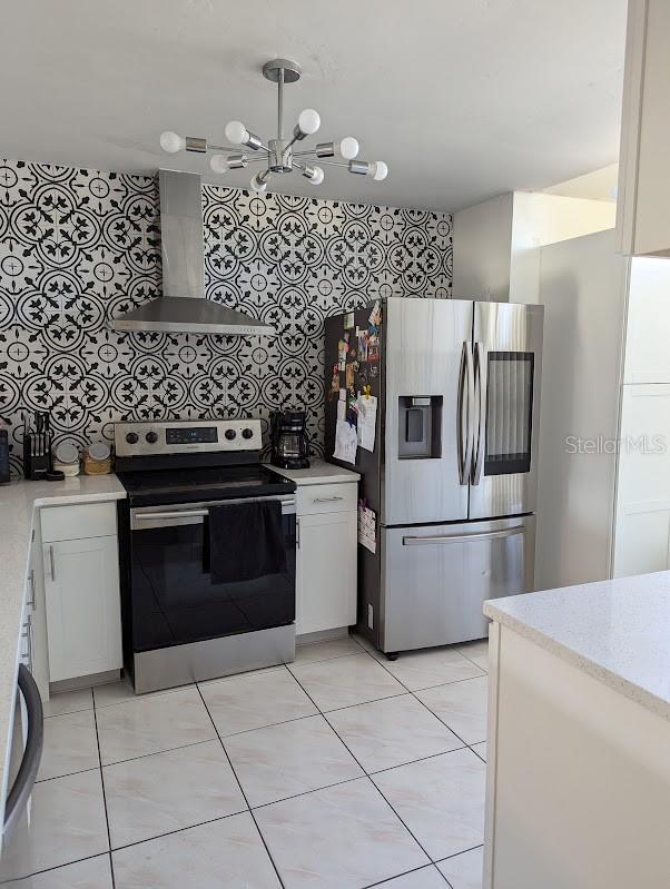 kitchen featuring decorative backsplash, stainless steel appliances, wall chimney range hood, a notable chandelier, and white cabinets