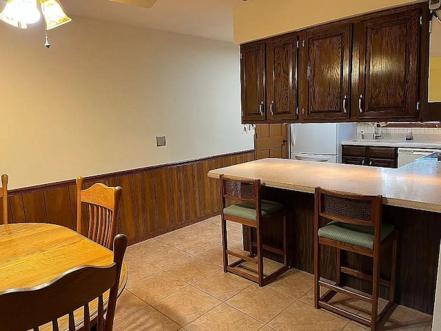 kitchen with dark brown cabinets, light tile patterned floors, dishwasher, white fridge, and wood walls