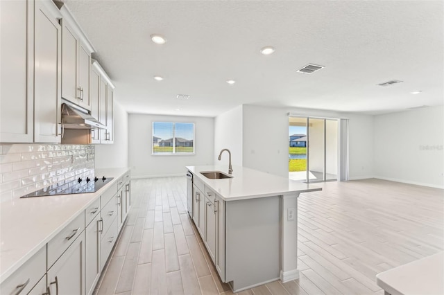 kitchen with an island with sink, black electric cooktop, sink, light hardwood / wood-style flooring, and backsplash