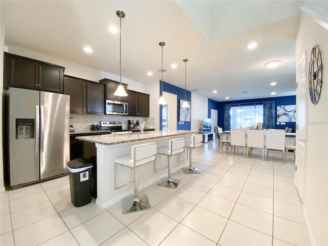 kitchen featuring a center island with sink, decorative light fixtures, light stone counters, dark brown cabinetry, and stainless steel appliances