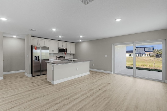 kitchen with a kitchen island with sink, dark stone counters, light wood-type flooring, white cabinetry, and stainless steel appliances