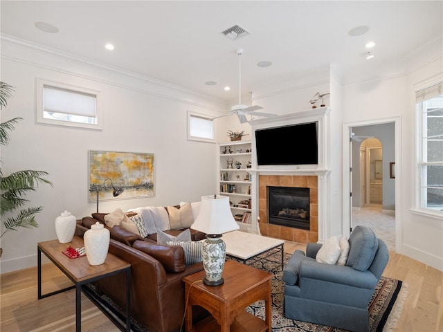 living room featuring built in shelves, ceiling fan, crown molding, a tile fireplace, and light hardwood / wood-style flooring