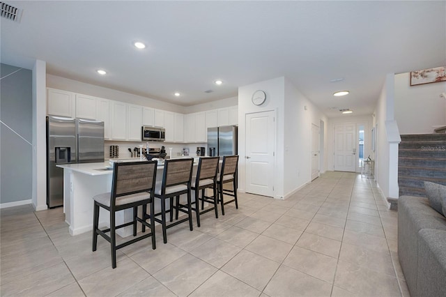 kitchen featuring a kitchen island with sink, white cabinets, light tile patterned floors, appliances with stainless steel finishes, and a breakfast bar area