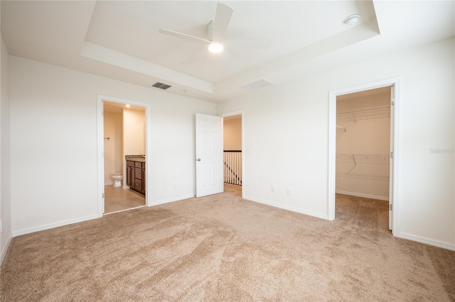 unfurnished bedroom with a spacious closet, ceiling fan, ensuite bathroom, a tray ceiling, and a closet