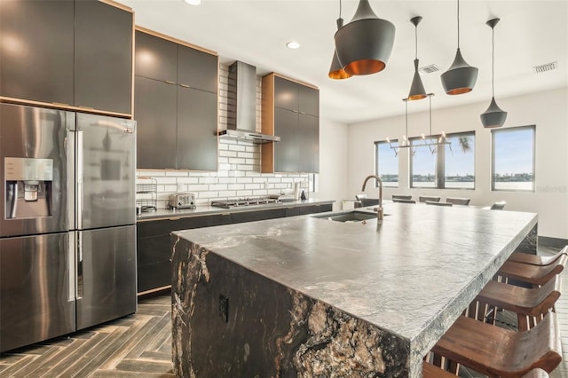 kitchen featuring a kitchen island with sink, hanging light fixtures, a breakfast bar area, wall chimney exhaust hood, and stainless steel appliances