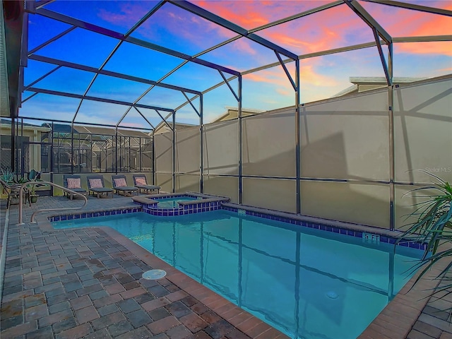 pool at dusk featuring glass enclosure, a patio area, and an in ground hot tub