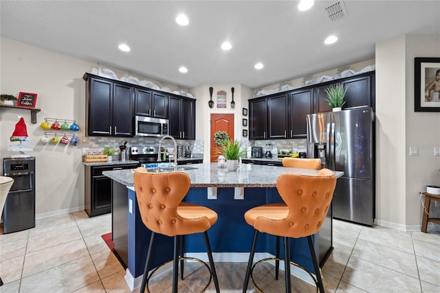 kitchen featuring a kitchen island with sink, a breakfast bar area, light stone countertops, light tile patterned flooring, and stainless steel appliances