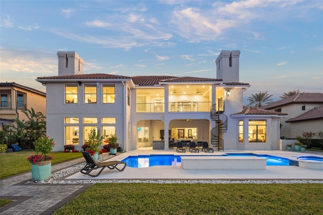 back house at dusk with a swimming pool with hot tub, a balcony, a yard, and a patio