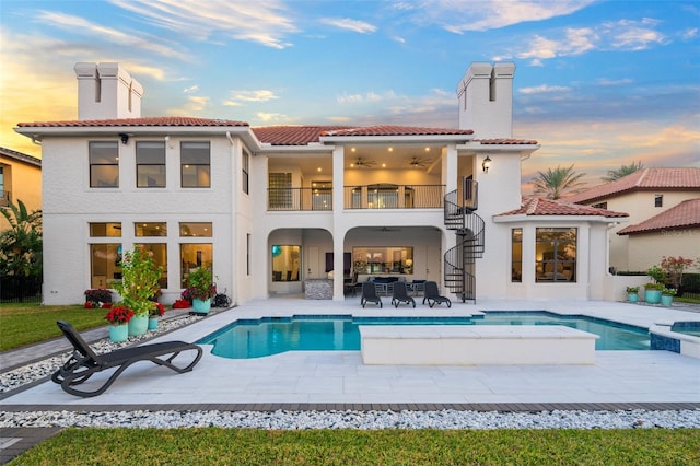 back house at dusk featuring a balcony, a swimming pool with hot tub, and a patio