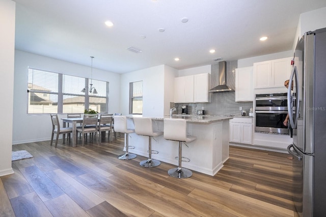 kitchen featuring stainless steel appliances, wall chimney range hood, pendant lighting, a kitchen island with sink, and white cabinets