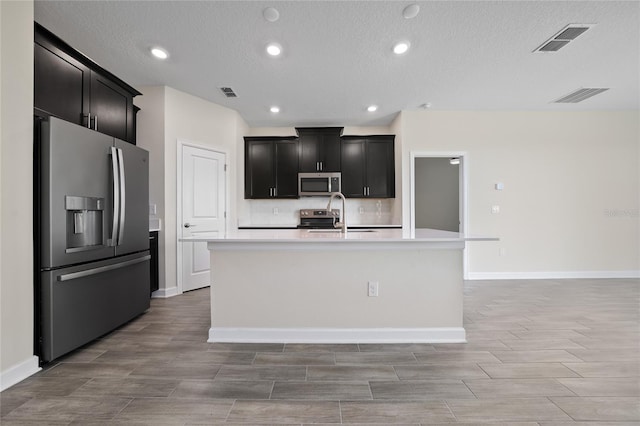 kitchen featuring appliances with stainless steel finishes, tasteful backsplash, a textured ceiling, sink, and a center island with sink