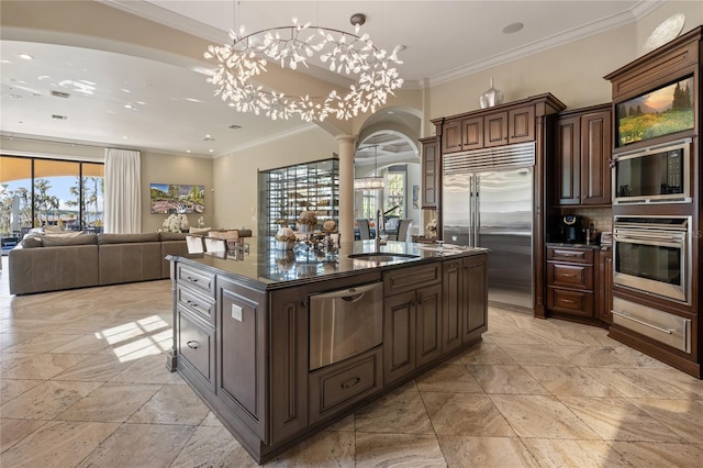 kitchen with a center island with sink, sink, built in appliances, ornate columns, and dark brown cabinetry