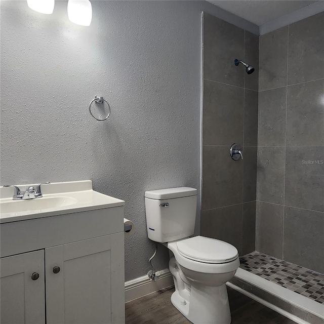 bathroom featuring toilet, wood finished floors, a tile shower, a textured wall, and vanity