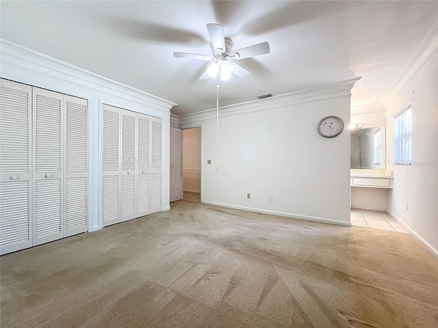 unfurnished bedroom featuring ensuite bath, ornamental molding, light colored carpet, ceiling fan, and multiple closets