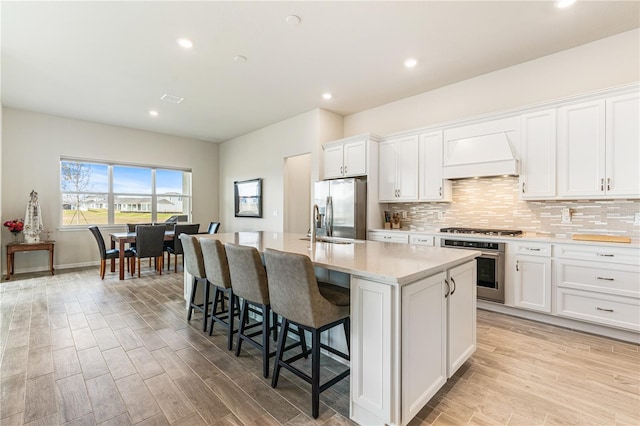 kitchen with premium range hood, a center island with sink, light wood-type flooring, appliances with stainless steel finishes, and white cabinetry