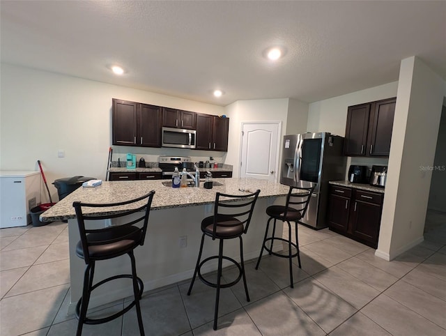 kitchen featuring a breakfast bar, a kitchen island with sink, light tile patterned floors, appliances with stainless steel finishes, and dark brown cabinets