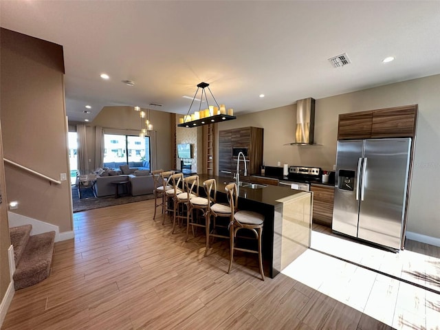 kitchen with appliances with stainless steel finishes, wall chimney exhaust hood, sink, pendant lighting, and a center island with sink