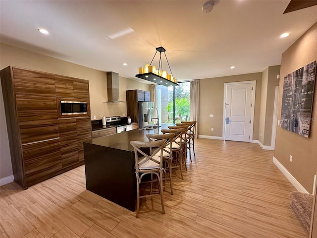 kitchen with appliances with stainless steel finishes, a kitchen breakfast bar, wall chimney range hood, decorative light fixtures, and light hardwood / wood-style flooring