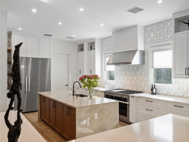 kitchen with a center island with sink, wall chimney range hood, light stone countertops, white cabinetry, and stainless steel appliances