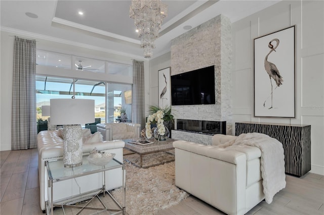 living room with ceiling fan with notable chandelier, ornamental molding, a raised ceiling, and a stone fireplace