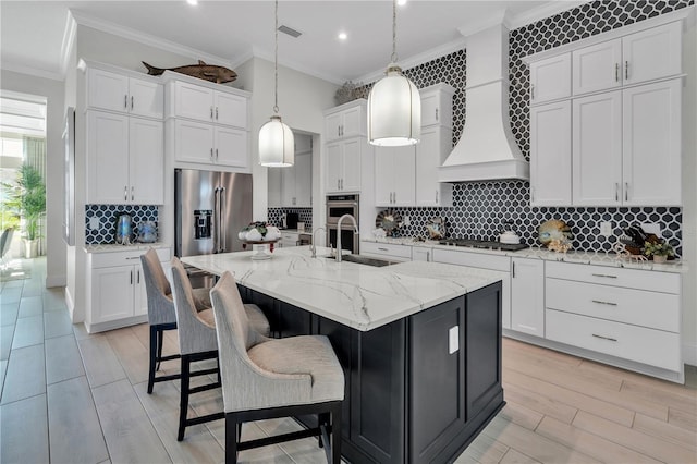 kitchen with premium range hood, hanging light fixtures, an island with sink, stainless steel appliances, and white cabinets