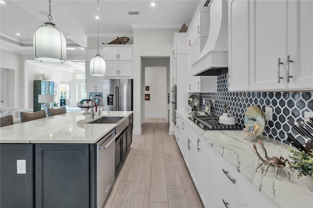 kitchen with custom exhaust hood, white cabinetry, stainless steel appliances, hanging light fixtures, and a kitchen island with sink