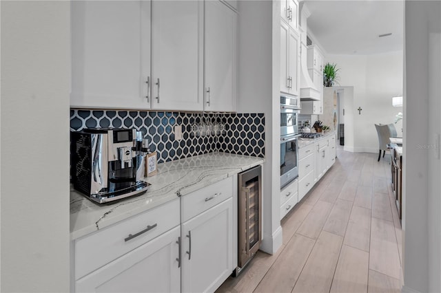 kitchen with light stone countertops, stainless steel double oven, beverage cooler, white cabinets, and backsplash