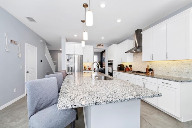 kitchen with a center island with sink, wall chimney exhaust hood, stainless steel appliances, and decorative light fixtures